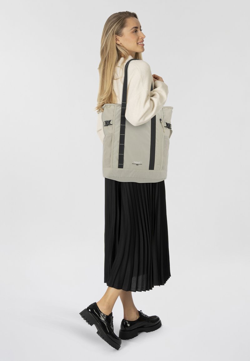 Borsa a spalla grigia chiara con manici neri, dotata di tasche con zip e una texture opaca e liscia. Il modello indossa una gonna nera plissettata e scarpe nere.