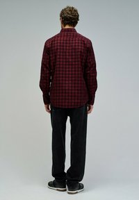 Mies, joka seisoo selin, päällä punamusta ruudullinen paita, mustat farkut ja mustat tennarit yksinkertaista harmaata taustaa vasten.