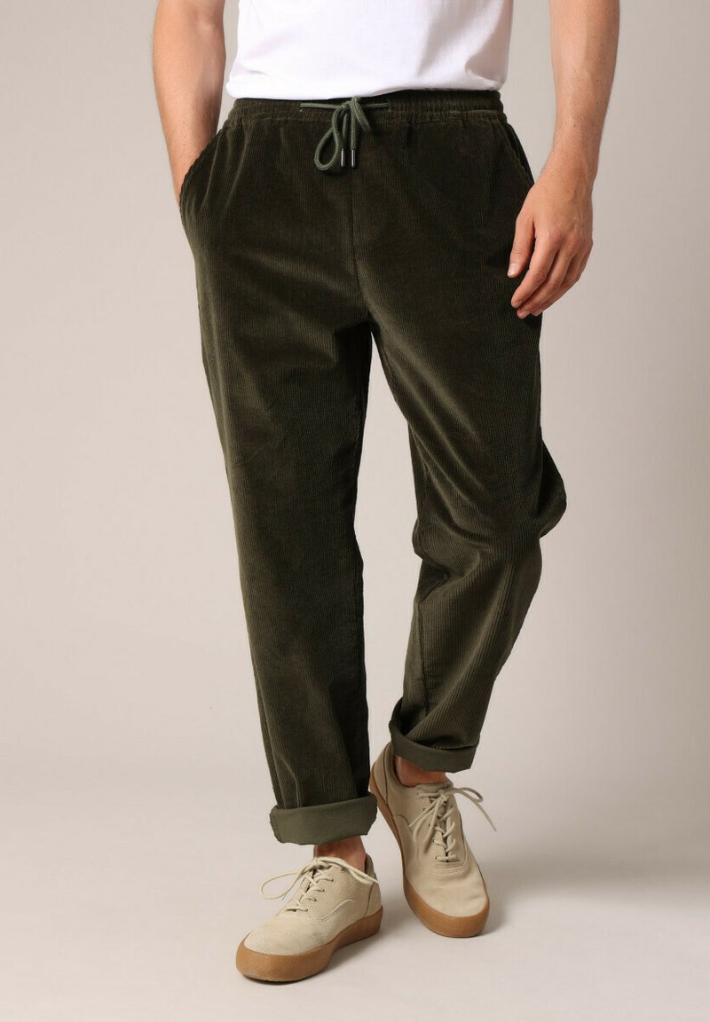 Olijfgroene corduroy broek met een elastische tailleband en verstelbaar trekkoord. Afgeronde uiteinden en zijzakken, gecombineerd met lichte sneakers.