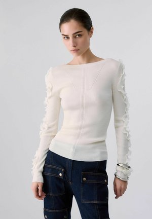 Femme aux cheveux foncés portant un pull en maille côtelée blanc avec des volants aux manches et un pantalon bleu marine avec de grandes poches avant.
