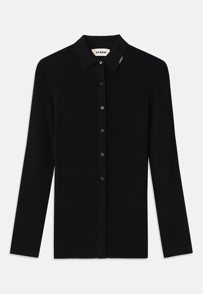 Cardigan lungo a maniche lunghe in rib cordonato nero con colletto a punta, chiusura frontale con bottoni e piccolo logo sul colletto. Texture liscia.