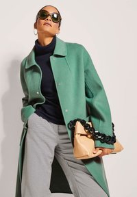 Groene wollen jas met een gekraagde halslijn, marineblauwe coltrui, grijs met patroon broek en een tan clutch met een zwarte kettinghandgreep.