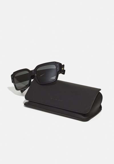 Ochelari de soare pătrați, de culoare neagră, cu lentile întunecate, decorati cu accente aurii și cu un finisaj mat, așezați lângă o carcasă din piele neagră.