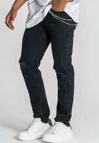 Jeans skinny negros con parches desgastados en las rodillas, mostrando una textura desvaída. Combinados con zapatillas blancas con detalles dorados.