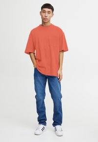 Camiseta de algodón naranja de corte relajado con cuello redondo y un bolsillo en el pecho, combinada con jeans de mezclilla azules y zapatillas blancas.