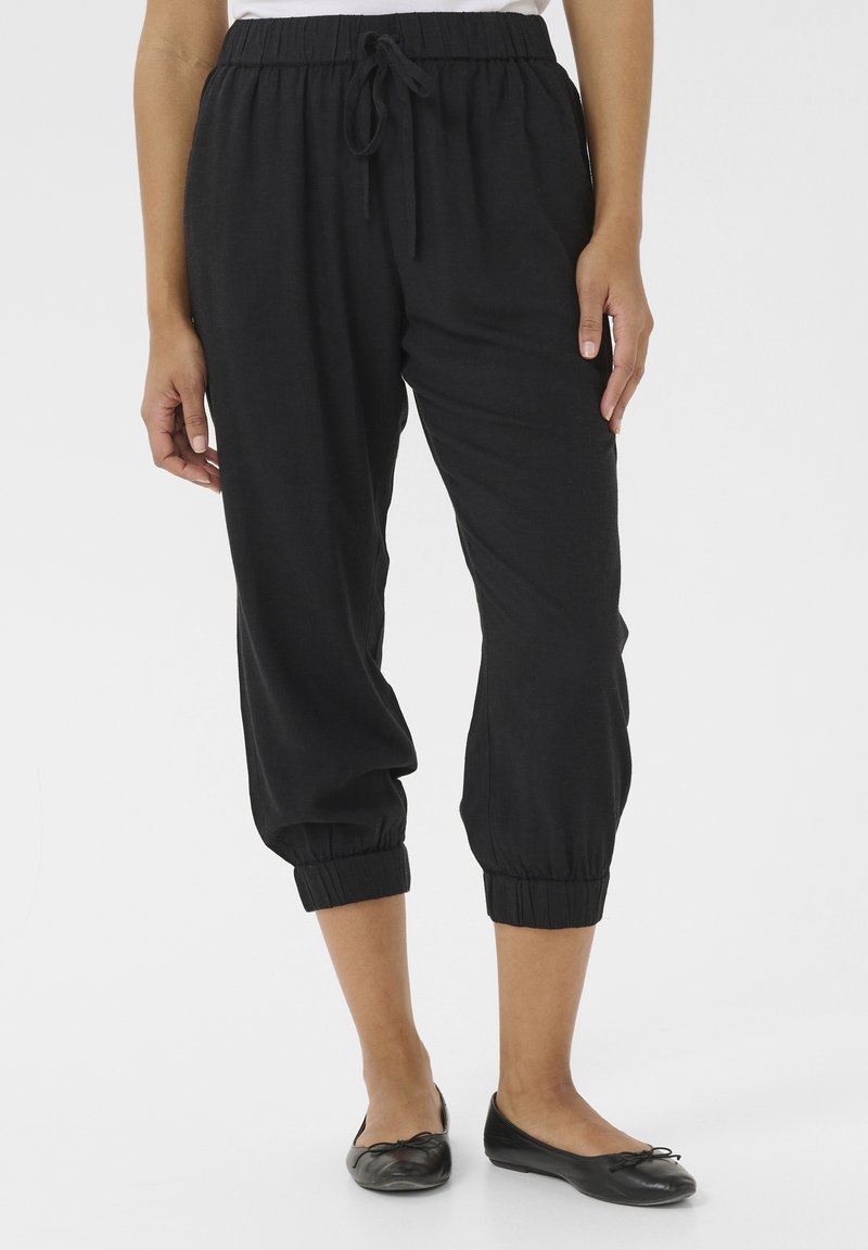 Žena v černých zkrácených jogger kalhotách s elastickým pasem a manžetami, obuta v černých balerínách, stojící proti bílému pozadí.