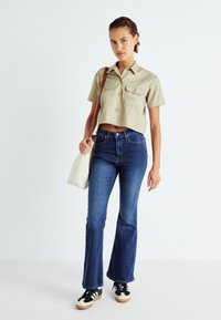 Camicia khaki tagliata con due tasche sul petto, abbinata a jeans blu svasati. Il modello porta una borsa a tracolla color crema e indossa sneakers nere.