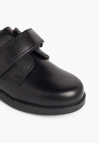 Chaussure en cuir noire avec un bout arrondi, une sangle en velcro et une semelle texturée avec des coutures visibles le long du bord.