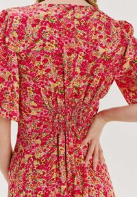 Červené květinové šaty s krátkými nabíranými rukávy, smockovaným pasem a živým vzorem růžových, žlutých a zelených květin.