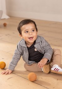 Gestreifte grau-weiße Jacke über einem schwarzen Hemd, beigen Hosen und weißen Sneakers mit roten Akzenten, sitzend auf einem Holzboden mit orangefarbenen Bällen.