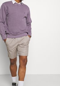 Felpa viola a girocollo con polsini a coste, indossata sopra una camicia bianca con colletto e pantaloni corti a maggio beige a scacchi, abbinata a calze bianche e scarpe nere.