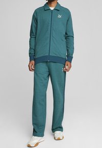 Teal tracksuit lavet af tekstureret stof, der har en zip-jakke og matchende bukser med marineblå accenter og et lille Puma-logo på brystet og benet.