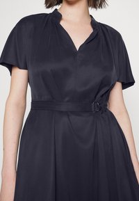 Mujer que lleva un vestido de manga corta azul oscuro con escote en V y una cintura ceñida con un detalle de hebilla lateral.