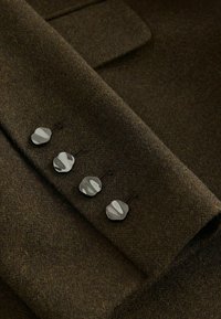Hnědá látka se čtyřmi lesklými knoflíky, které naznačují manžetu upraveného saka nebo kabátu. Je viditelná měkká textura.