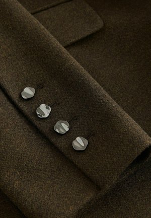 Tessuto di lana marrone con una texture morbida, caratterizzato da quattro bottoni argentati lucidi e curvi sul polsino per un dettaglio aggiuntivo e la chiusura.