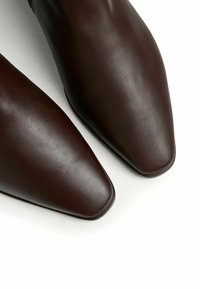 Botas de cuero marrón hasta el tobillo con punta afilada y textura suave. La parte superior tiene un diseño simple, y las suelas son planas y redondeadas.