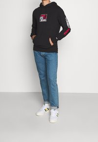 Zwarte katoenen hoodie met een frontgrafieklogo, lange mouwen met logodetail en een kangoeroezak, gecombineerd met lichtblauwe spijkerbroek.