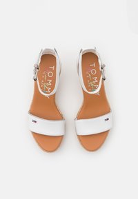 Bílé sandály na podpatku se širokým řemínkem, opálenou stélkou a espadrilovým stylem podrážky. Mají kovovou přezku a malý logo akcent.