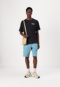 Black t-shirt with logo, blue shorts, beige crossbody bag, gray socks, and beige sneakers with accents. Smooth textures, casual design.