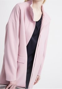 Light pink long coat with a high collar, side pockets, and a zip front closure, made of textured fabric. Worn over a dark top.