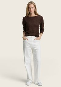 Pull en tricot marron avec un col large, associé à un pantalon en denim blanc taille haute. Le modèle se tient les mains dans les poches, portant des baskets blanches.