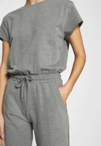 Personne portant un haut gris à manches courtes et un pantalon gris à cordon assorti, main dans la poche, sur un fond clair uni.