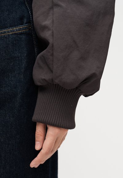 Χέρι χαλαρό στο πλάι, φορώντας μανίκι σακακιού με σκουρόχρωμη ριμπ ύφανση και σκουρόχρωμο τζιν με εμφανείς ραφές.
