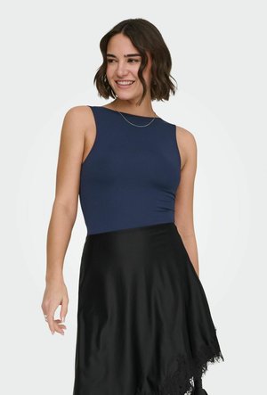 Kobieta o krótkich brązowych włosach, ubrana w granatowy top na ramiączkach i czarną spódnicę z koronkową lamówką, uśmiechająca się i patrząca w bok.