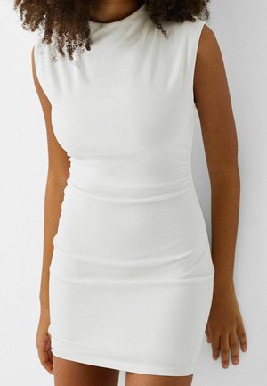 Femme aux cheveux bouclés portant une mini robe blanche ajustée sans manches, debout devant un fond blanc uni.