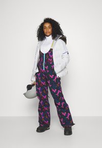 Schneeoverall mit einem schwarzen Blumenmuster in Pink und Lila. Kombiniert mit einem weißen Rollkragenpullover und einer weißen Daunenjacke. Schwarze Stiefel und ein Helm.