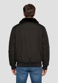 Giacca bomber nera con colletto di pelliccia, caratterizzata da una finitura liscia, polsini ed elastico in vita. Indossata con jeans blu, mostrando una vestibilità casual.