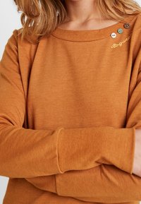 Un pull couleur rouille avec un décolleté large, comportant des boutons décoratifs et un texte brodé sur l'épaule. Tissu doux et texturé.