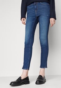 Jeans blu slim fit indossati con mocassini in pelle nera e una maglia scura a maniche lunghe, mostrati dalla metà della coscia fino ai piedi su uno sfondo semplice.