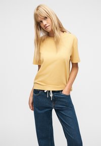 Ξανθιά γυναίκα με ίσια μαλλιά φοράει κίτρινη μπλούζα με κοντά μανίκια και μπλε τζιν, στέκεται με το ένα χέρι στην τσέπη, μπροστά σε λευκό φόντο.
