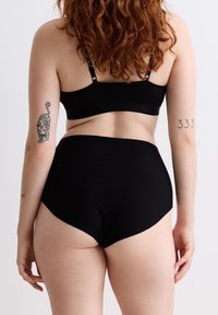 Frau mit lockigem rotem Haar, die schwarze Unterwäsche trägt und ein Leopard-Tattoo am linken Arm sowie "333" am rechten Arm hat, von hinten gesehen.