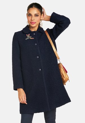 Marineblauer texturierter Mantel mit Kragen, fünf Knöpfen und seitlichen Taschen, ausgestattet mit einer bunten Blumenbrosche und kombiniert mit einer braunen Handtasche.