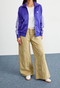 Person som bär en lila Adidas träningsjacka, vit kort topp, vida beige cargobyxor med dragkedjor och vita sneakers på blå matta.