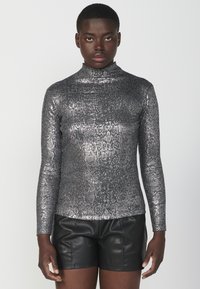 Metallic srebrna dugosvjetlosna majica s visokim ovratnikom, koja ima teksturirani uzorak zmijskin printom. Uparena s crnim kožnim šortsevima.