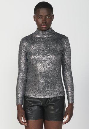 Metallisch silbernes Langarmoberteil mit Stehkragen, das ein strukturiertes Schlangenprintdesign aufweist. Kombiniert mit schwarzen Ledershorts.