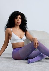 Λιλά καλσόν με λαμπερό, γκλίτερ υφή, σε αποχρώσεις του μοβ, σε συνδυασμό με λευκό μπράλετ. Το μοντέλο κάθεται σε ένα μαλακό, ουδέτερο χαλί.