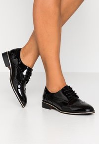 Černé lakované kožené boty s nízkým podpatkem, zdobené brogue detaily a šněrováním, s lesklým povrchem a elegantním designem.