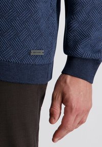 Navyblauer strukturierter Pullover mit Fischgratmuster, gerippten Bündchen und einem kleinen silbernen Markenanhänger nahe dem Saum.