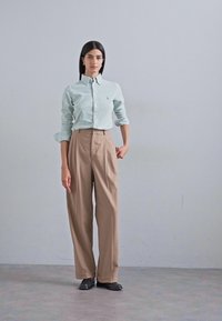 Camicia a righe blu chiaro e bianca con bottoni e maniche lunghe, abbinata a pantaloni beigi a vita alta. Scarpe nere completano l'outfit.