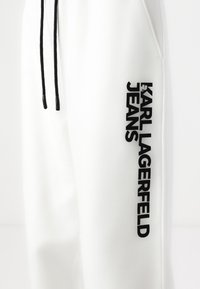 Baltos sportinės kelnės, pagamintos iš lygaus audinio, su juodomis raištelėmis ir ryškiu juodu logotipu šone, kuriame užrašyta "KARL LAGERFELD JEANS."