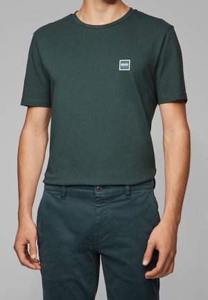 Mand iført mørkegrøn T-shirt med korte ærmer og matchende mørkegrønne bukser foran en ensfarvet grå baggrund.