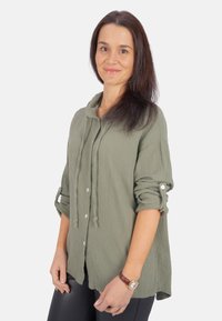 Grünes Button-Up-Hemd mit Kapuze, strukturierter Stoff, hochgerollte Ärmel und lockere Passform. Braue Uhr am Handgelenk.