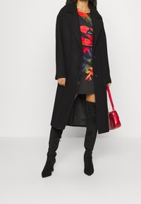 Un manteau en laine noir avec un col cranté, comportant deux poches avant. Associé à une robe à motifs floraux, des bottes noires montantes et un sac à main rouge.