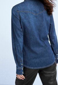 Denim shirt in dark blue with long sleeves, buttoned cuffs, and a yoke design on the back; paired with black pants.
