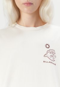 Cream-colored cotton t-shirt featuring a brown cartoon wave graphic and sun design, with "BILLABONG" text below the graphic on the left chest.