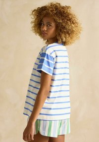 Svītrainā T-kreklā baltā un gaiši zilā krāsā, īsām piedurknēm, brīvs piegriezums. Ievērojams zīmols uz piedurknes. Zaļi un zili svītrainas šortus.